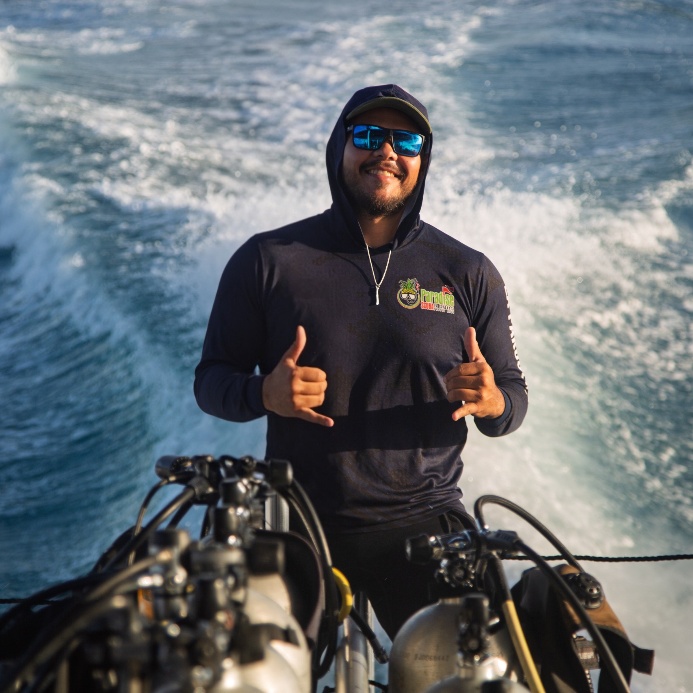 Person on a boat giving a thumbs-up gesture with scuba tanks in the foreground.