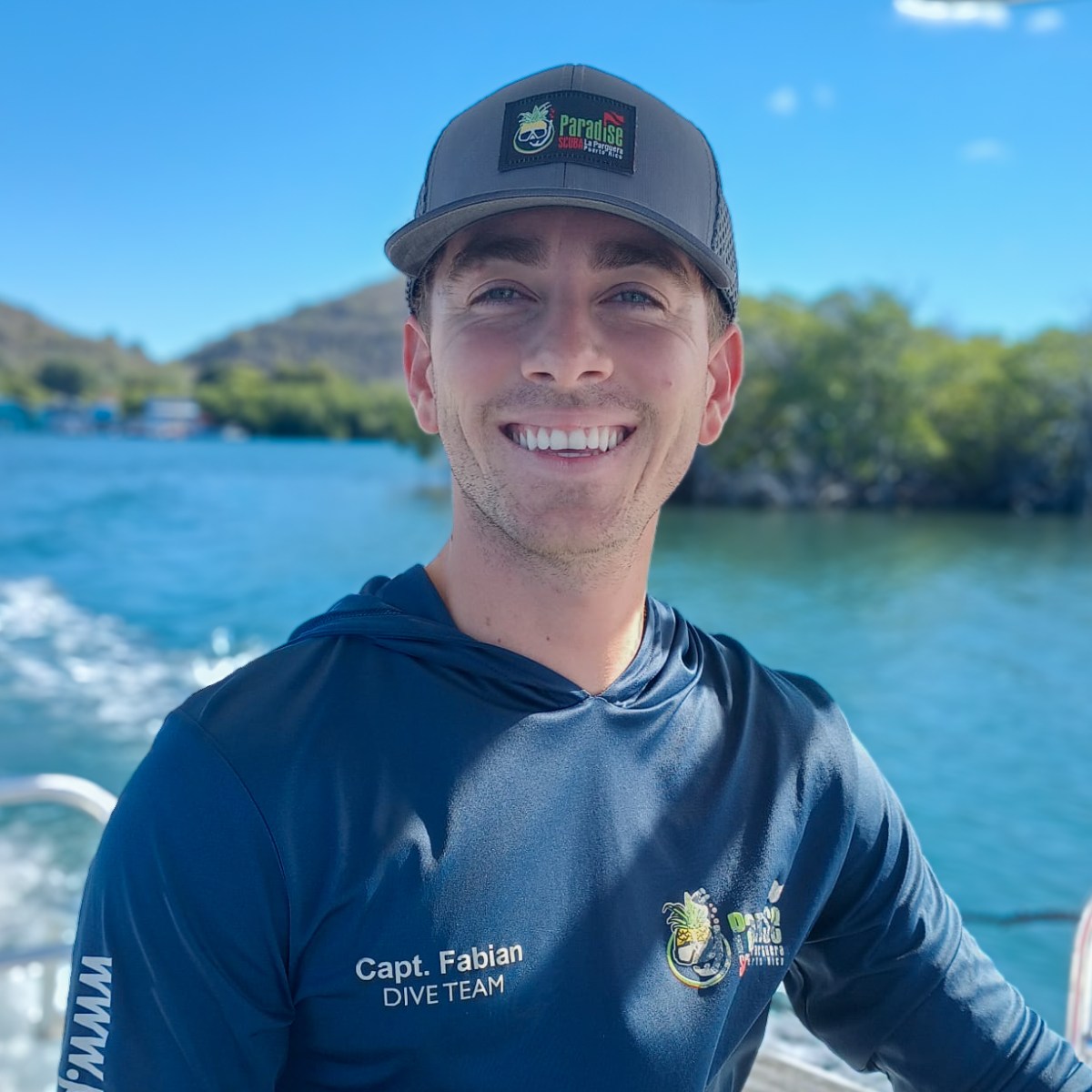 Smiling man in a cap on a boat by water and green hills.