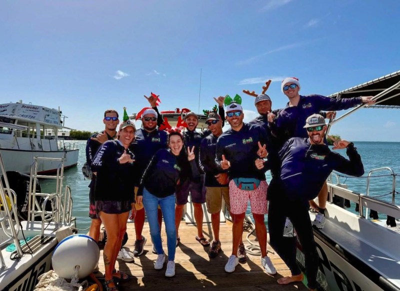 Group of 11 people posing on dock with boats, wearing festive hats and casual clothes.