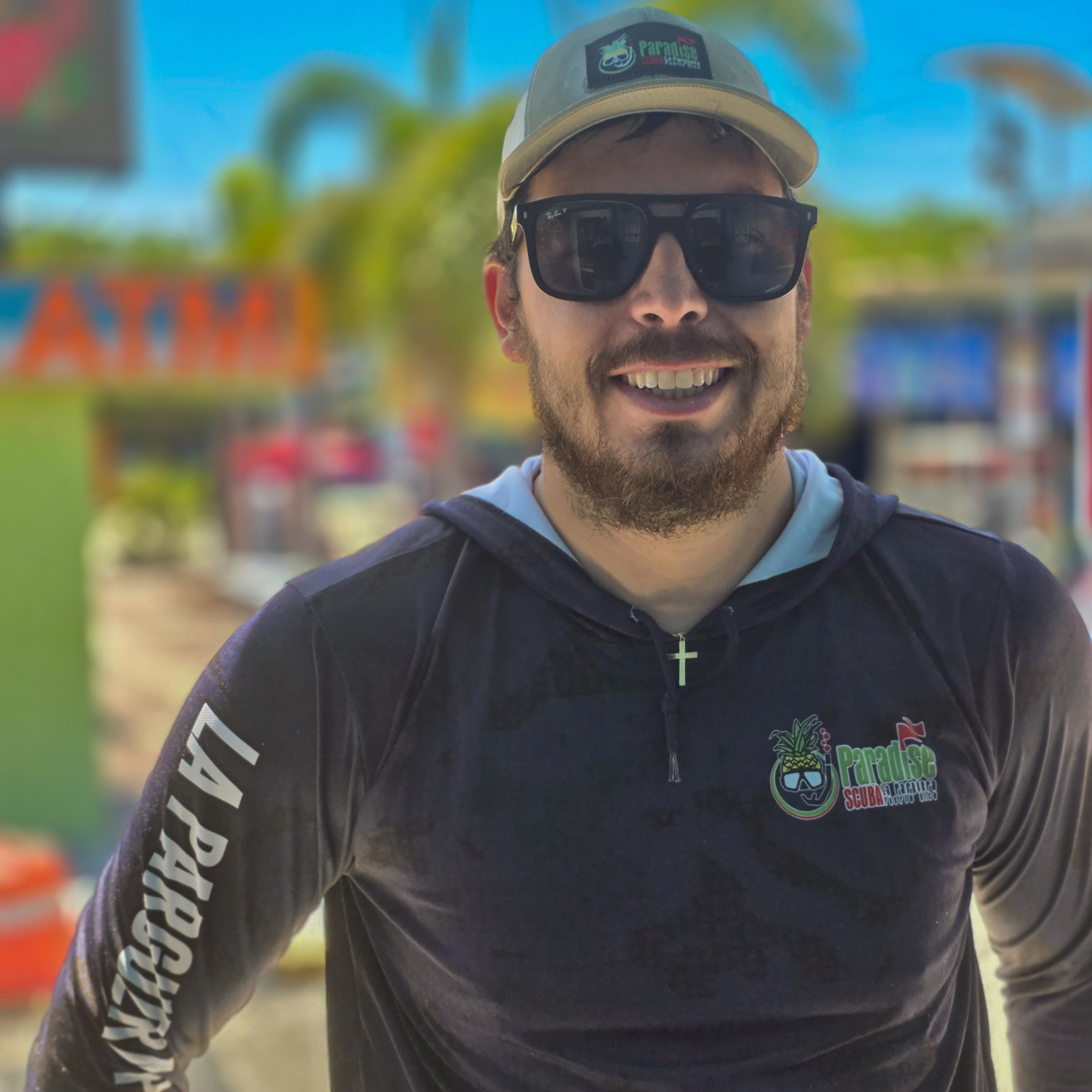 Man with sunglasses and cap smiling outdoors in sunny weather.