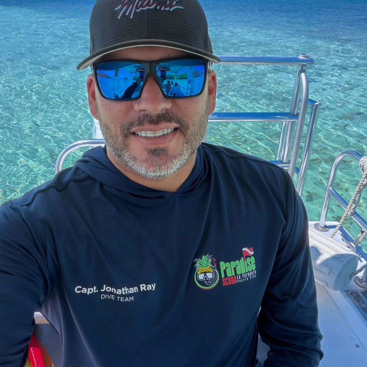 Man in sunglasses and cap on a boat with clear blue water and hills in background.